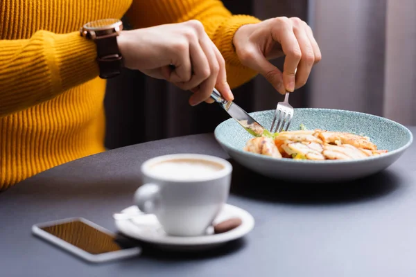 Vista ritagliata dell'uomo che taglia l'insalata vicino a smartphone e tazza di cappuccino in primo piano sfocato — Foto stock