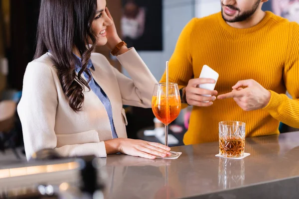 Smiling woman looking at friend pointing at smartphone near beverages on bar counter on blurred foreground — Stock Photo
