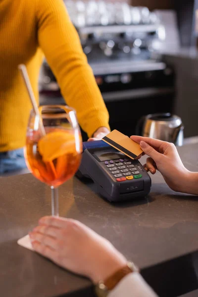 Vista cortada de mulher segurando cartão de crédito perto do terminal de pagamento, bartender e coquetel em primeiro plano turvo — Fotografia de Stock