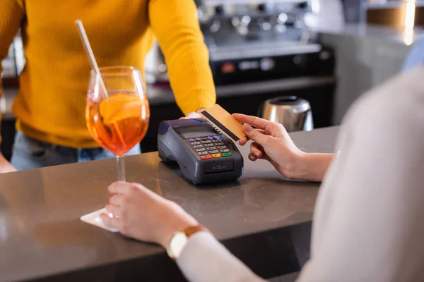 Vue recadrée de la femme payant avec carte de crédit près du barman et cocktail au premier plan flou — Photo de stock