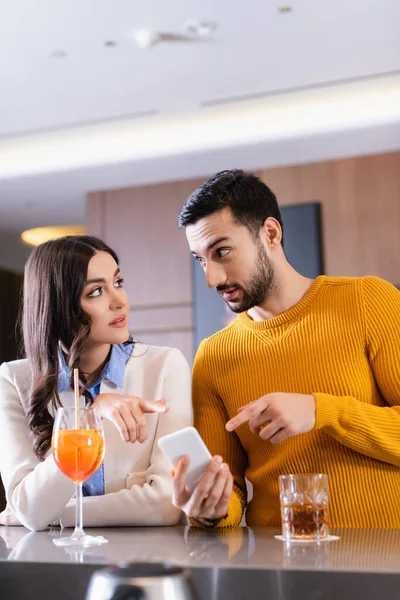 Woman pointing at smartphone near muslim friend and drinks on blurred foreground in restaurant — Stock Photo