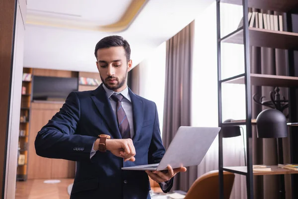 Uomo d'affari musulmano guardando orologio da polso e tenendo il computer portatile nel ristorante — Foto stock