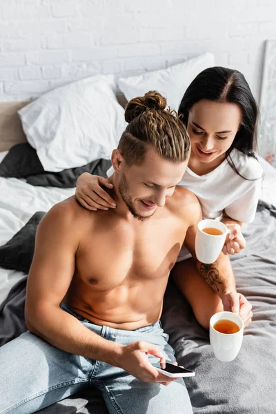 Shirtless man using smartphone near girlfriend with cup on bed — Stock Photo