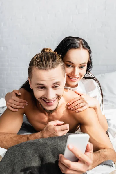 Cheerful man using smartphone near girlfriend in bedroom — Stock Photo