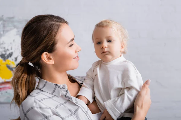 Happy young mother looking at little son while holding him at home — Stock Photo