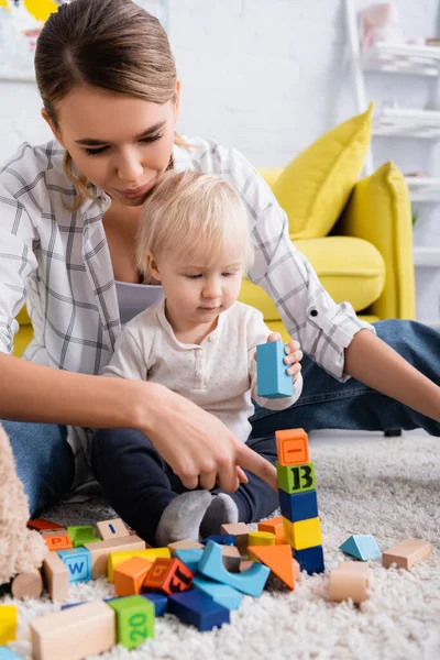 Young woman pointing with finger at colorful cubes while playing with son on carpet, blurred foreground — Stock Photo