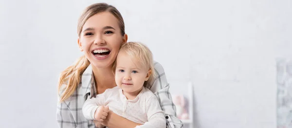 Excited young woman looking at camera while holding smiling child, banner — Stock Photo