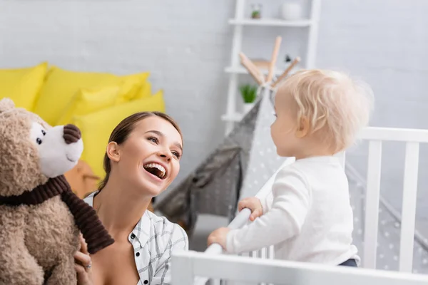 Excited woman holding teddy bear near little son standing in crib — Stock Photo