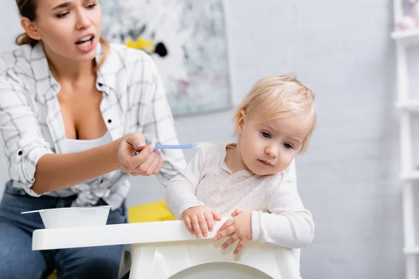 Young mother feeding little son turning away from spoon while sitting on kids chair, blurred background — Stock Photo