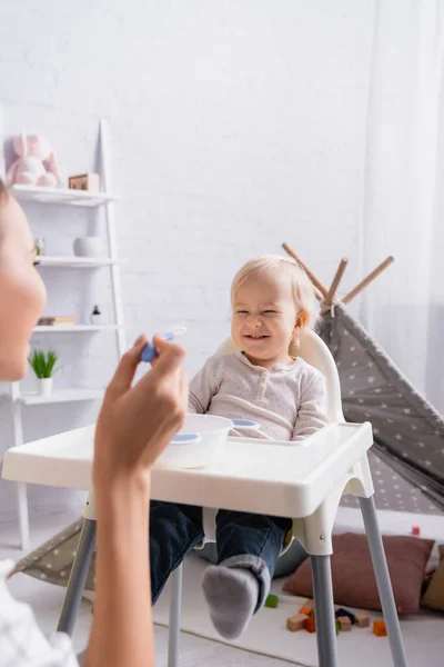 Woman holding spoon near cheerful baby boy sitting on kids chair, blurred foreground — Stock Photo