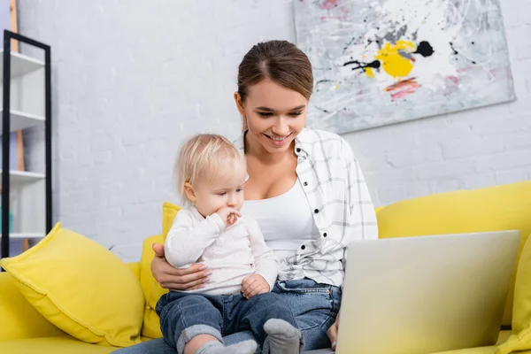 Smiling freelancer using laptop while sitting on yellow sofa with little son — Stock Photo