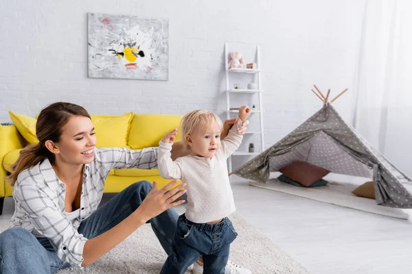 Sonriente madre ayudando a su pequeño hijo a caminar en el suelo en casa - foto de stock