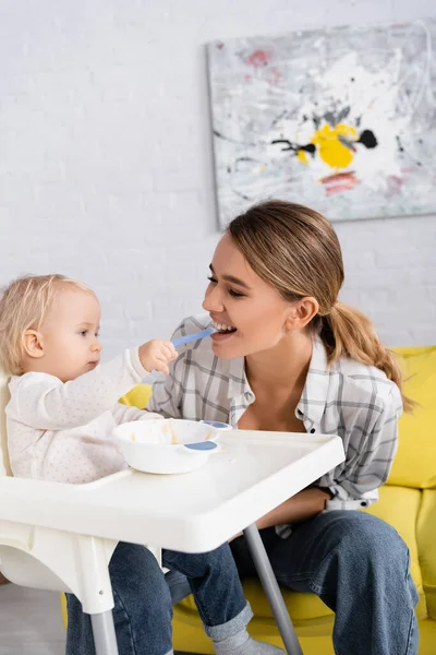 Baby boy sitting on kids chair and feeding cheerful mother — Stock Photo