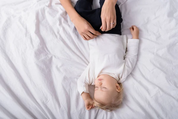 Top view of mother buttoning romper of little son sleeping on white bedding — Stock Photo