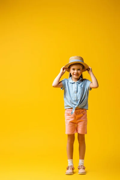 Full length of happy child adjusting straw hat on yellow — Stock Photo