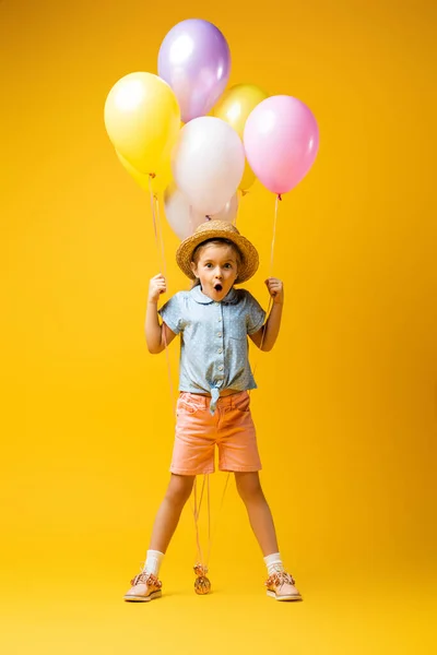 Full length of shocked kid in straw hat holding balloons on yellow — Stock Photo