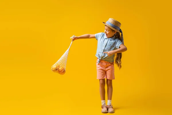Longitud completa de niño excitado en sombrero de paja que apunta con el dedo en el bolso de cadena reutilizable con naranjas en amarillo - foto de stock