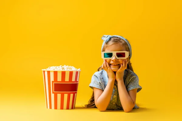 Glückliches Kind in 3D-Gläsern liegt neben Popcorn-Eimer auf gelb — Stockfoto