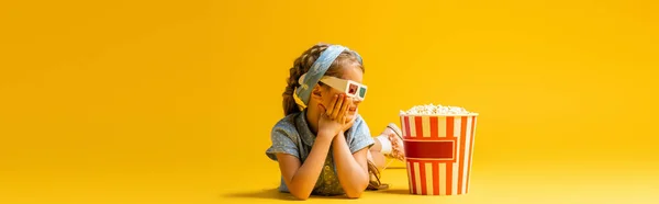 Happy kid in 3d glasses lying near popcorn bucket on yellow, banner — Stock Photo