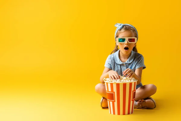 Niño sorprendido en gafas 3D sentado con las piernas cruzadas cerca de palomitas de maíz cubo y viendo la película en amarillo - foto de stock