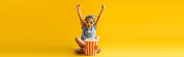 Excited kid in 3d glasses sitting with crossed legs and raised hands near popcorn bucket and watching movie on yellow, banner — Stock Photo