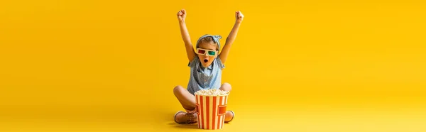 Enfant excité dans des lunettes 3D assis avec les jambes croisées et les mains levées près du seau de maïs soufflé et regarder le film sur jaune, bannière — Photo de stock