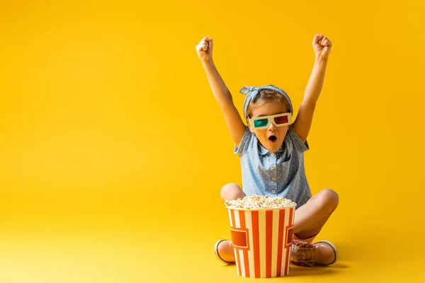 Excited kid in 3d glasses sitting with crossed legs and raised hands near popcorn bucket and watching movie on yellow — Stock Photo