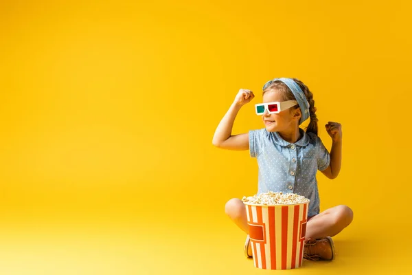 Niño sonriente en gafas 3D sentado con las piernas cruzadas cerca de palomitas de maíz cubo en amarillo - foto de stock