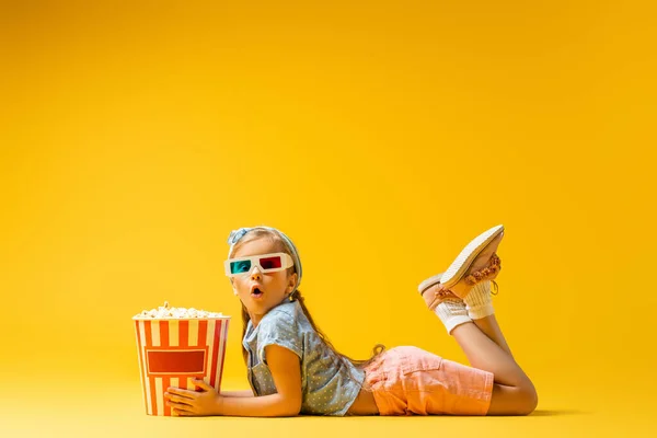 Chocado chica en 3d gafas acostado cerca palomitas de maíz cubo en amarillo — Stock Photo