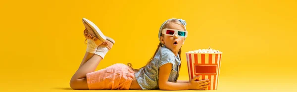 Shocked girl in 3d glasses lying near popcorn bucket on yellow, banner — Stock Photo