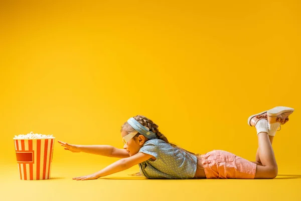 Vista lateral de niño en gafas 3d acostado y alcanzando palomitas de maíz cubo en amarillo — Stock Photo