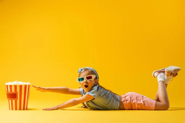 Überraschtes Mädchen in 3D-Brille liegt und greift nach Popcorn-Eimer auf gelb — Stockfoto