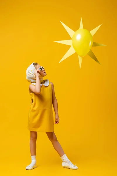 Pleine longueur de fille en foulard et lunettes de soleil regardant le soleil décoratif avec ballon sur jaune — Photo de stock