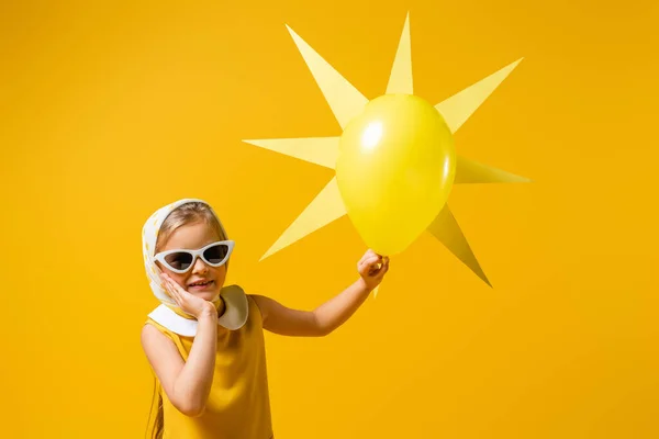 Happy girl in headscarf and sunglasses near decorative sun with balloon isolated on yellow — Stock Photo