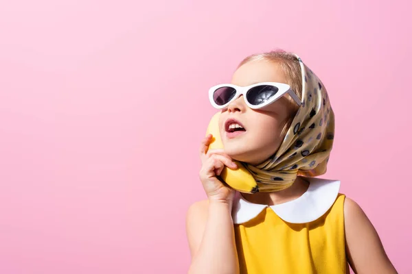 Niña en pañuelo para la cabeza y gafas de sol sosteniendo plátano cerca de la oreja aislado en rosa - foto de stock