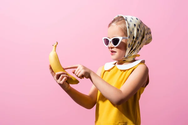 Chica feliz en pañuelo para la cabeza y gafas de sol apuntando con el dedo al plátano aislado en rosa - foto de stock