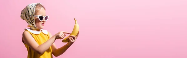 Счастливая девушка в платке и солнцезащитных очках указывая пальцем на банан изолирован на розовый, баннер — стоковое фото