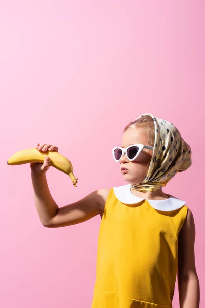 Girl in headscarf and sunglasses puffing cheeks while holding banana isolated on pink — Stock Photo
