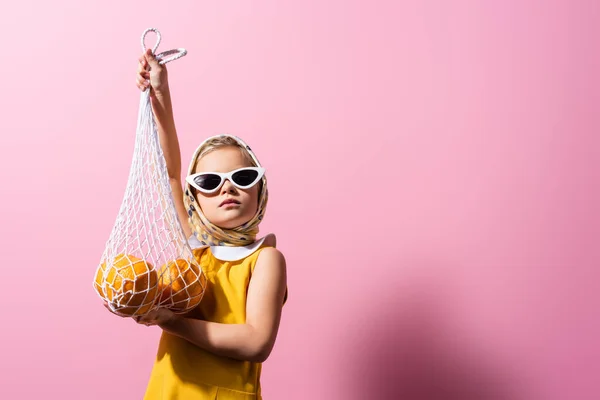 Kid in headscarf and sunglasses holding reusable string bag with oranges on pink — Stock Photo