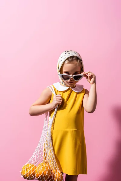 Kid in headscarf adjusting sunglasses while holding reusable string bag with oranges on pink — Stock Photo