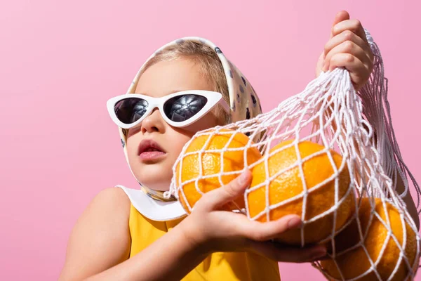 Elegante chica en pañuelo para la cabeza y gafas de sol con bolsa de hilo reutilizable con naranjas aisladas en rosa - foto de stock