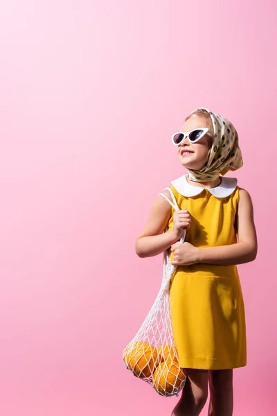 Alegre chica en pañuelo para la cabeza y gafas de sol sosteniendo bolsa de hilo reutilizable con naranjas aisladas en rosa - foto de stock