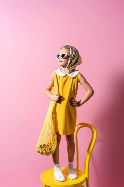 Cheerful girl in headscarf and sunglasses holding reusable string bag with bananas while standing on yellow chair on pink — Stock Photo