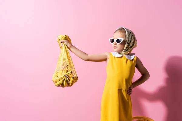 Niña en pañuelo para la cabeza y gafas de sol sosteniendo bolsa de hilo reutilizable con plátanos mientras está de pie con la mano en la cadera en rosa - foto de stock