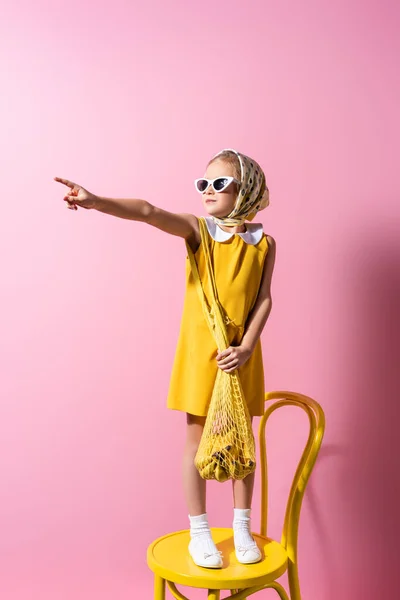 Fille en foulard et lunettes de soleil tenant sac à ficelle réutilisable avec des bananes tout en se tenant debout sur la chaise et pointant du doigt sur rose — Photo de stock