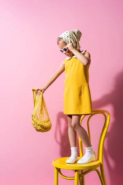Child in headscarf adjusting sunglasses while holding reusable string bag with bananas and standing on chair on pink — Stock Photo