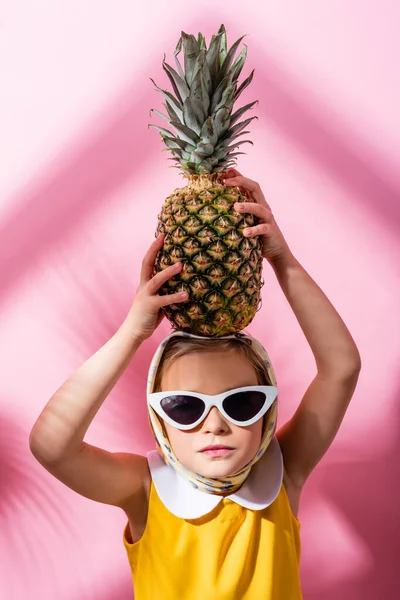 Mädchen mit Kopftuch und Sonnenbrille hält Ananas über dem Kopf auf rosa — Stockfoto