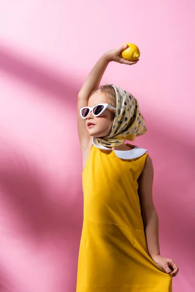 Chica con estilo en el pañuelo para la cabeza y gafas de sol con limón por encima de la cabeza en rosa - foto de stock