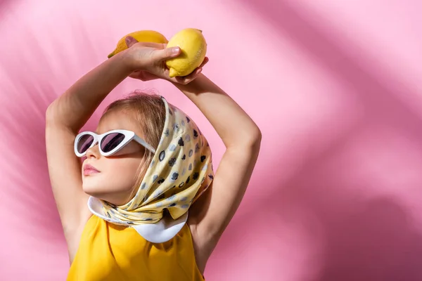 Chica con estilo en pañuelo para la cabeza y gafas de sol celebración de limones por encima de la cabeza en rosa - foto de stock