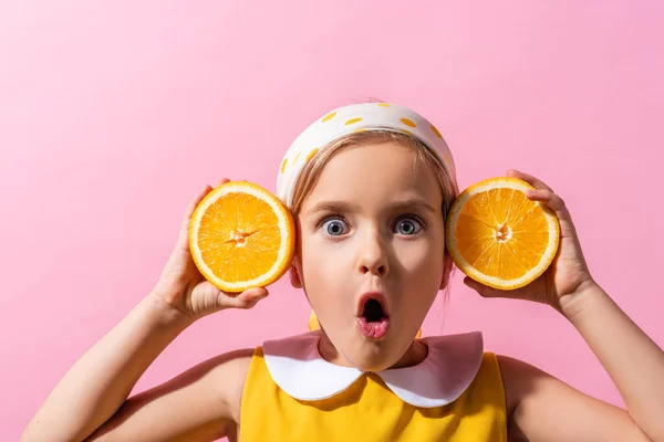 Surprised girl in headscarf holding orange halves near ears isolated on pink — Stock Photo
